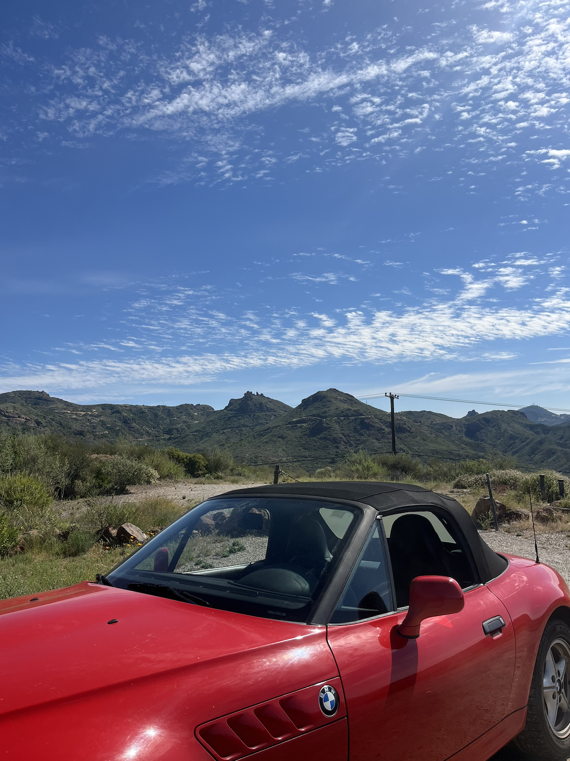 A red BMW convertible car parked in a rural area with green hills and a blue sky with white clouds in the background.
