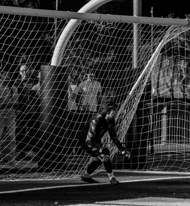 Youth soccer goalkeeper in a black uniform crouches in front of the goal during a match, with a torn goal net, onlookers in the background.