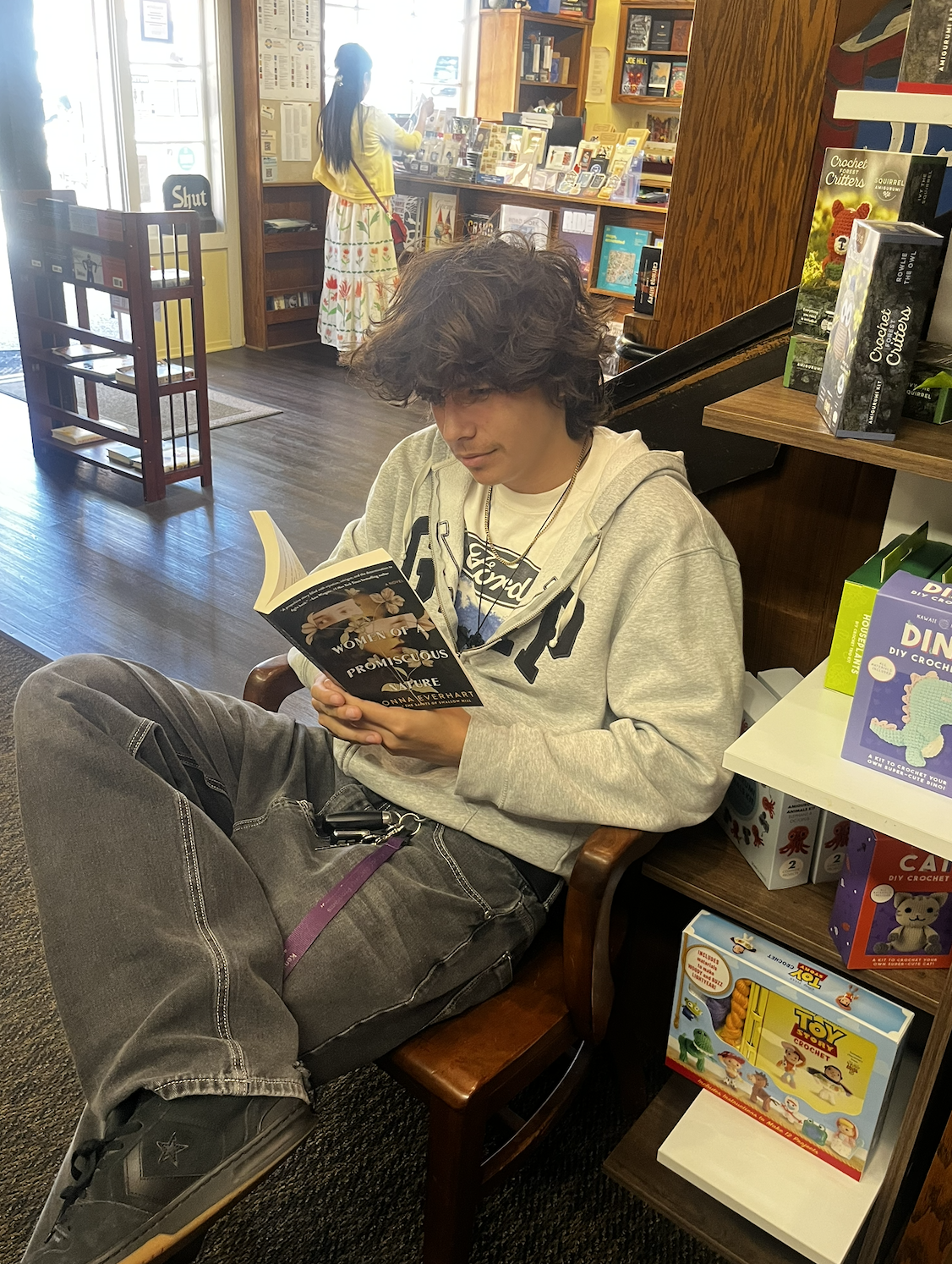 A person with curly hair reading a book in a bookstore, sitting in a wooden chair with legs crossed, wearing a gray hoodie, black pants, and black sneakers.