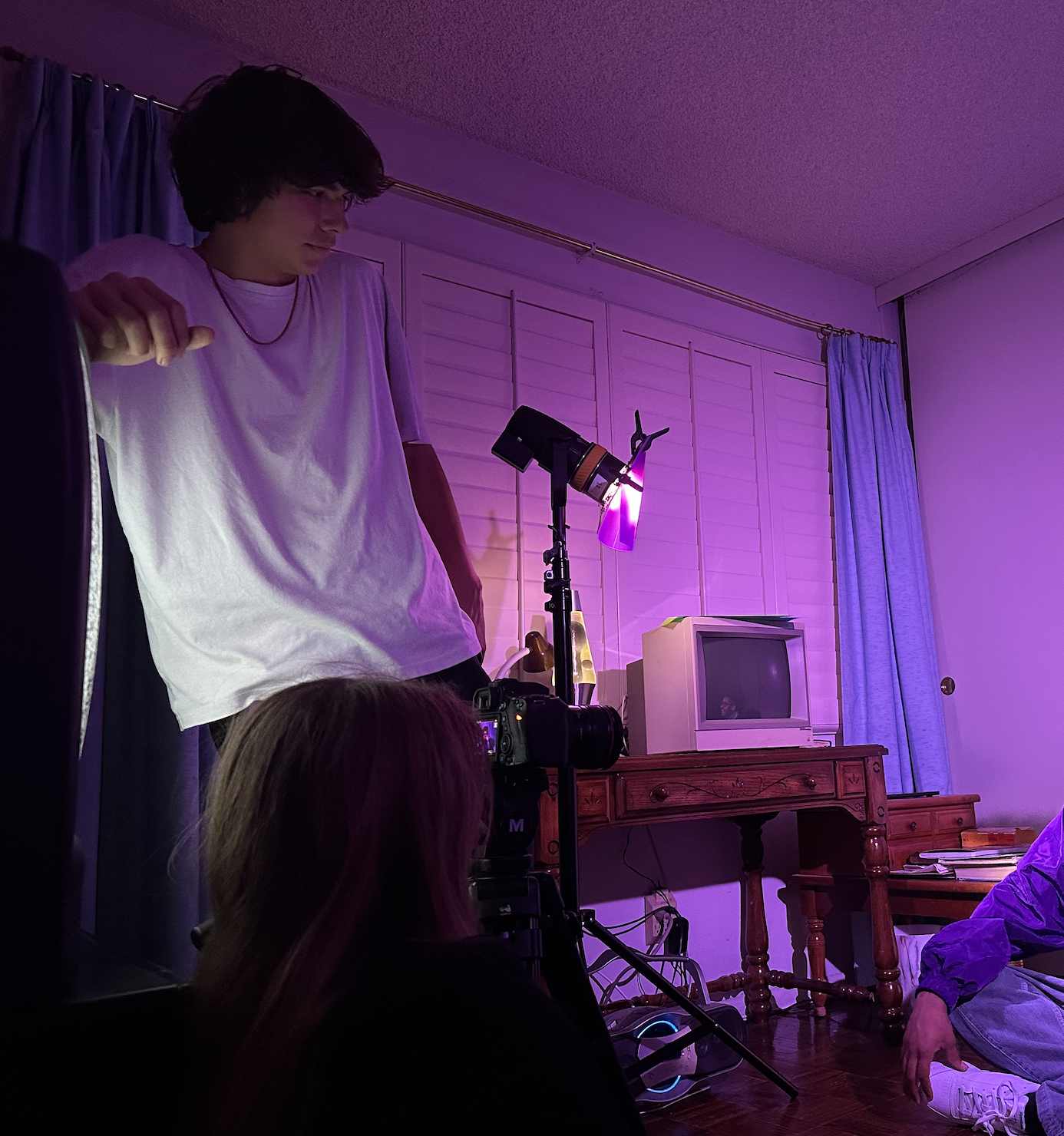 Young man standing in a room with purple lighting, next to a camera on a tripod, with a small TV and a wooden cabinet in the background, and people seated on the floor.