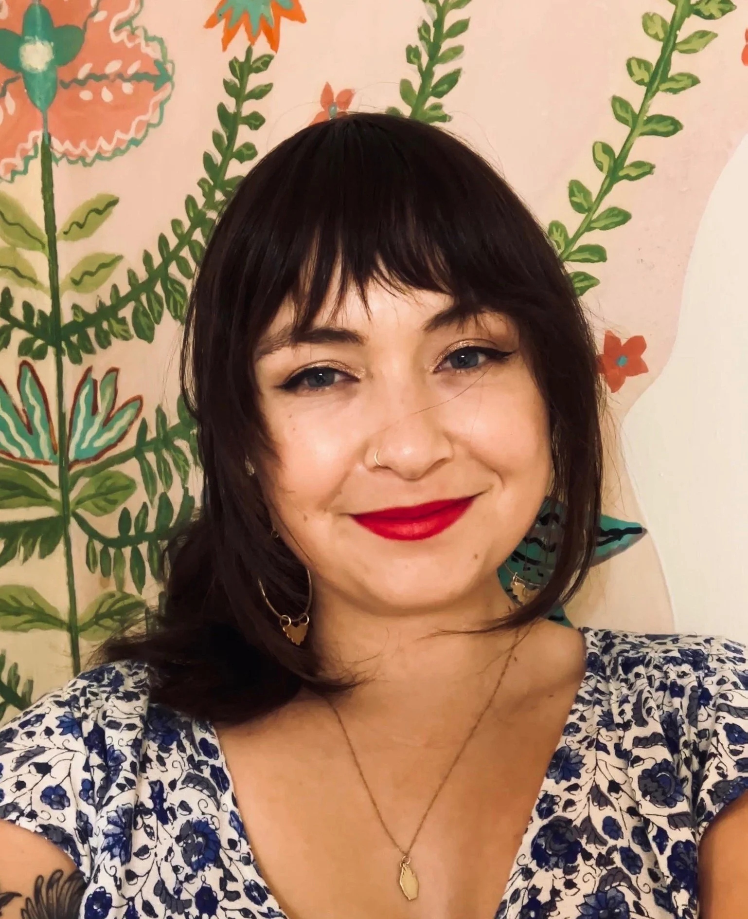 Close-up of a smiling woman with dark hair, red lipstick, and gold earrings, wearing a floral patterned top and a gold pendant necklace, with a colorful botanical mural background.