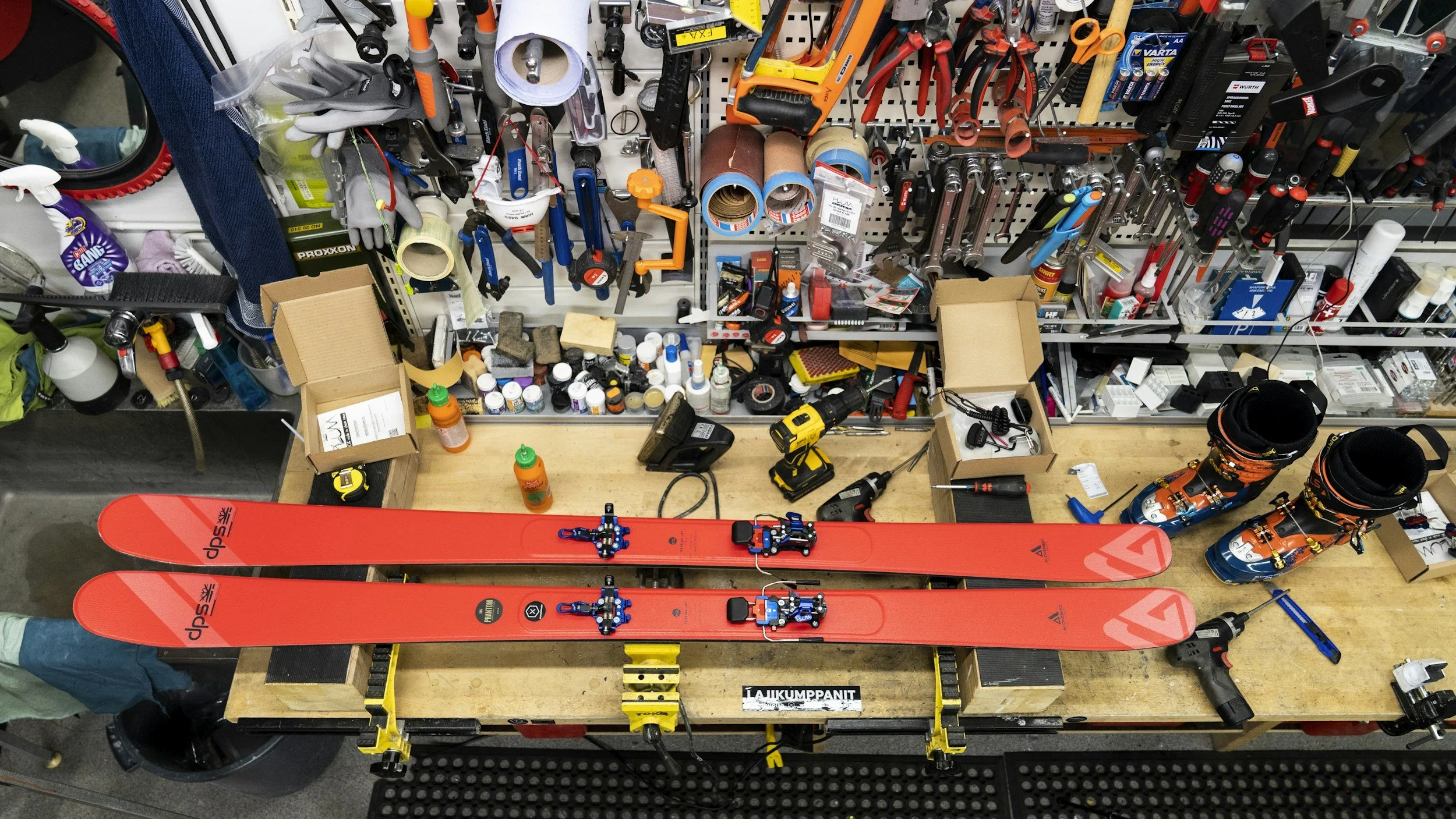 Image of a cluttered workbench with ski equipment, tools, and supplies. Two red skis with bindings are at the front, with various tools, tape, and bottles behind them. There are orange and black ski boots at the side, and a pegboard above holding gloves, tape, screwdrivers, and other small items.