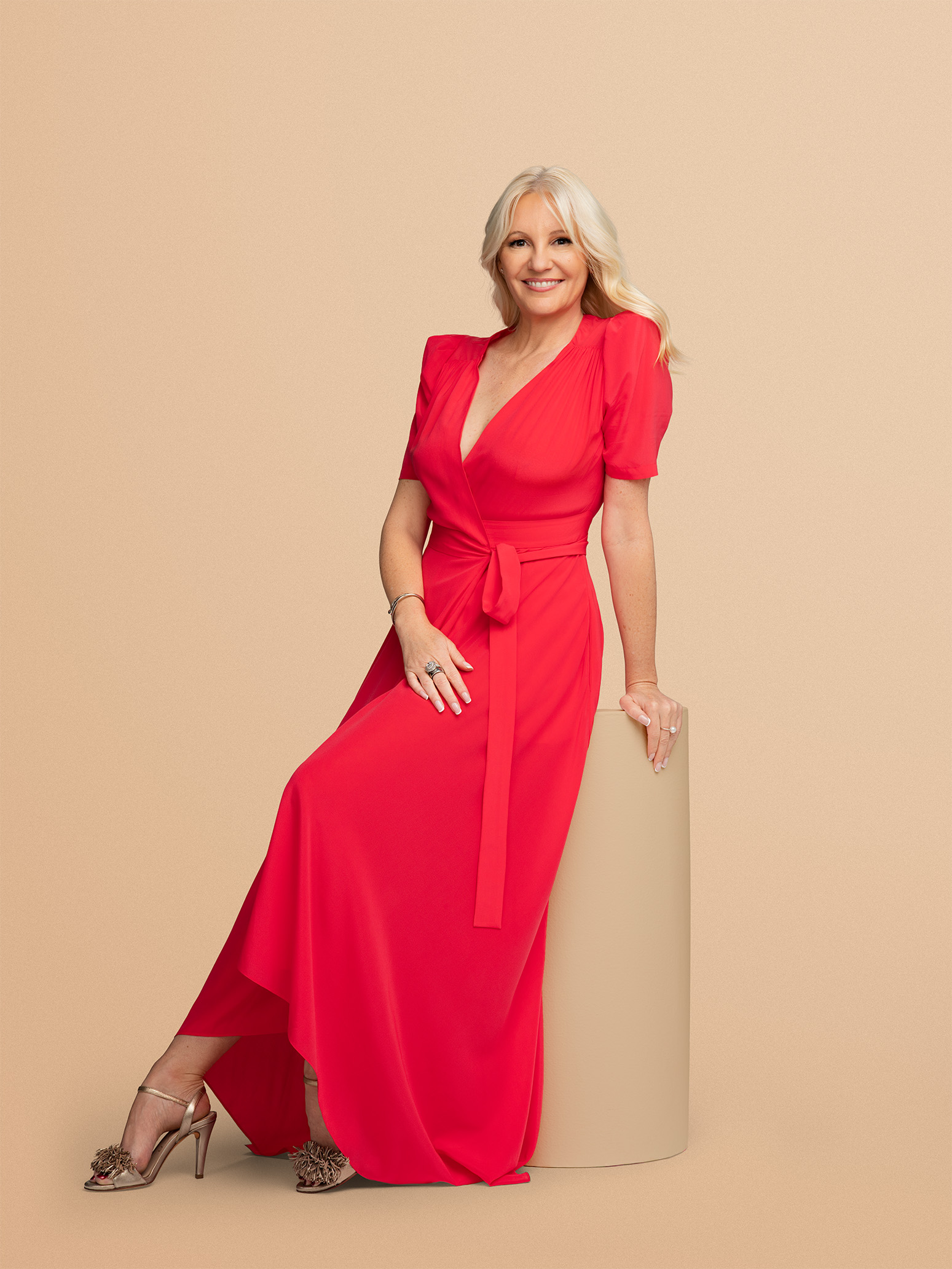 A woman in a red dress sitting on a beige cylindrical object against a beige background, wearing high heels with fluffy embellishments.