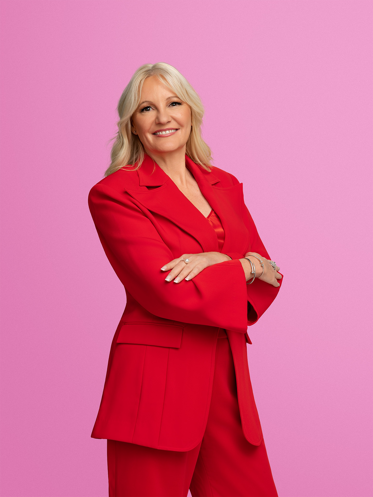 A woman with blonde hair smiling, wearing a red suit with crossed arms, standing against a pink background.