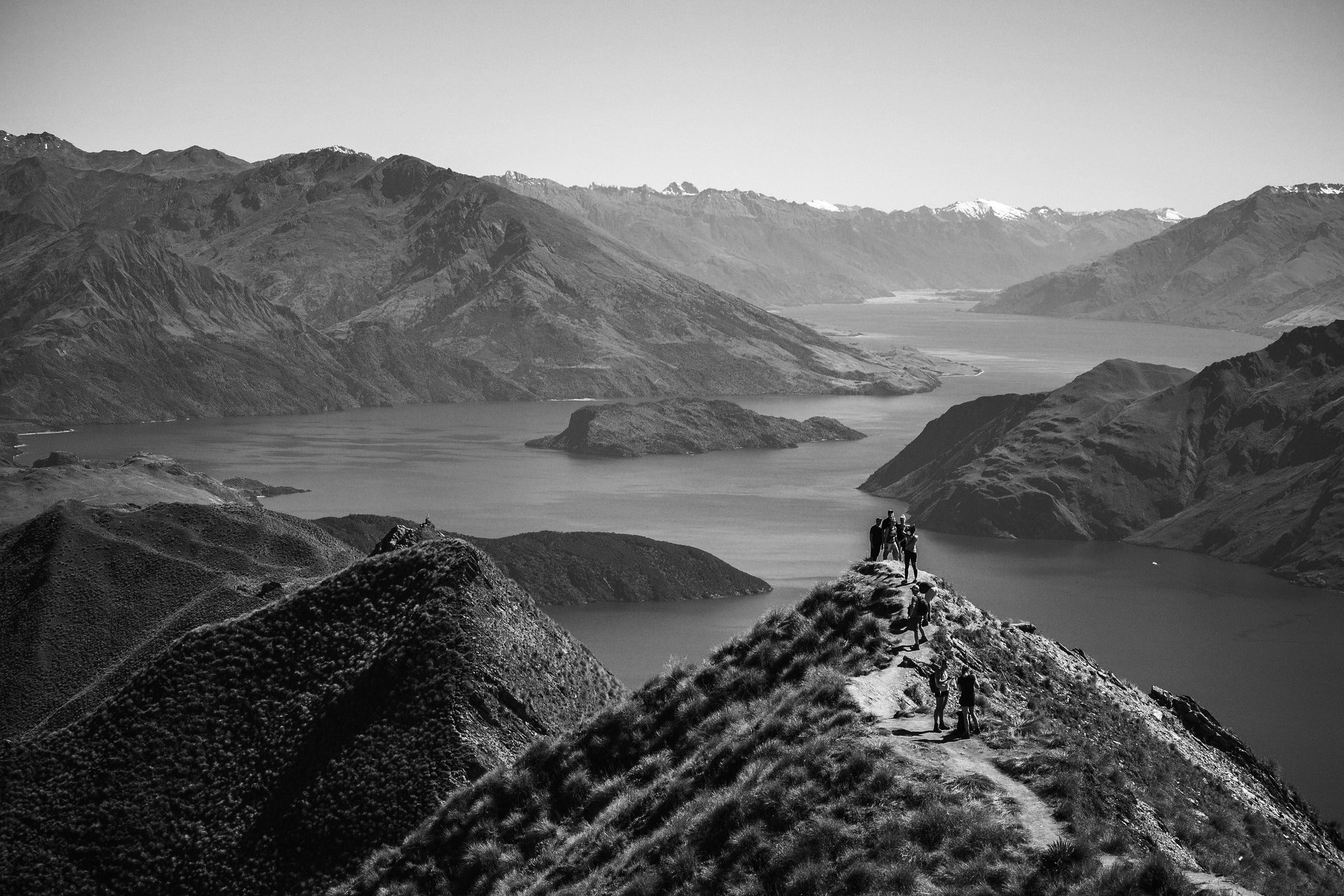 Black and white photo of hikers on a mountain ridge overlooking a lake, with mountains in the background.