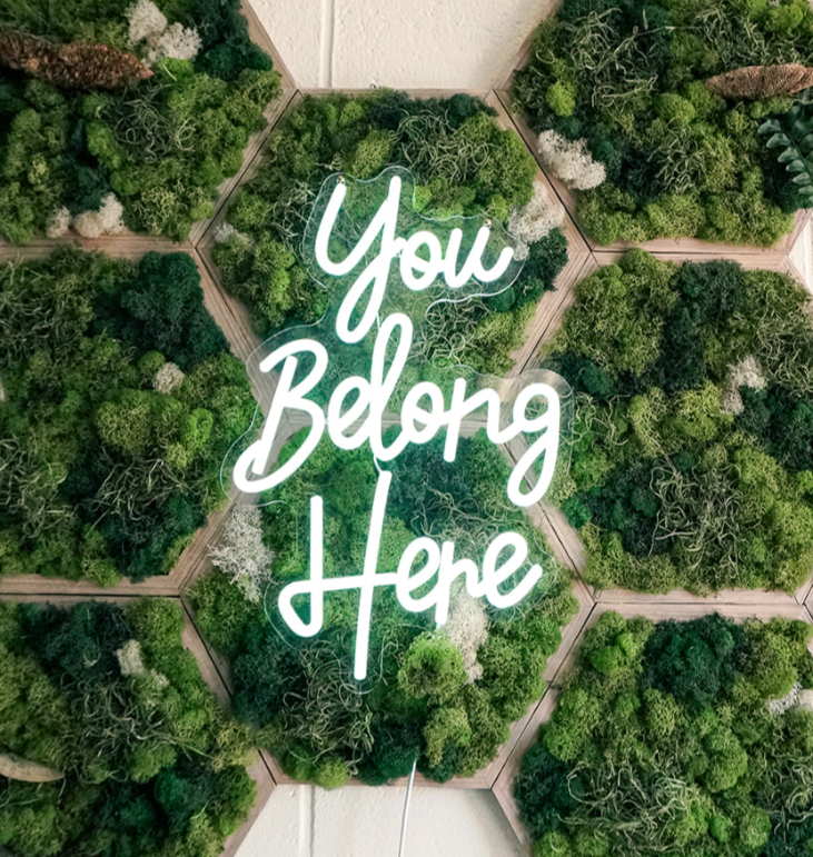 you belong here written in white neon on top of a honeycomb moss wall