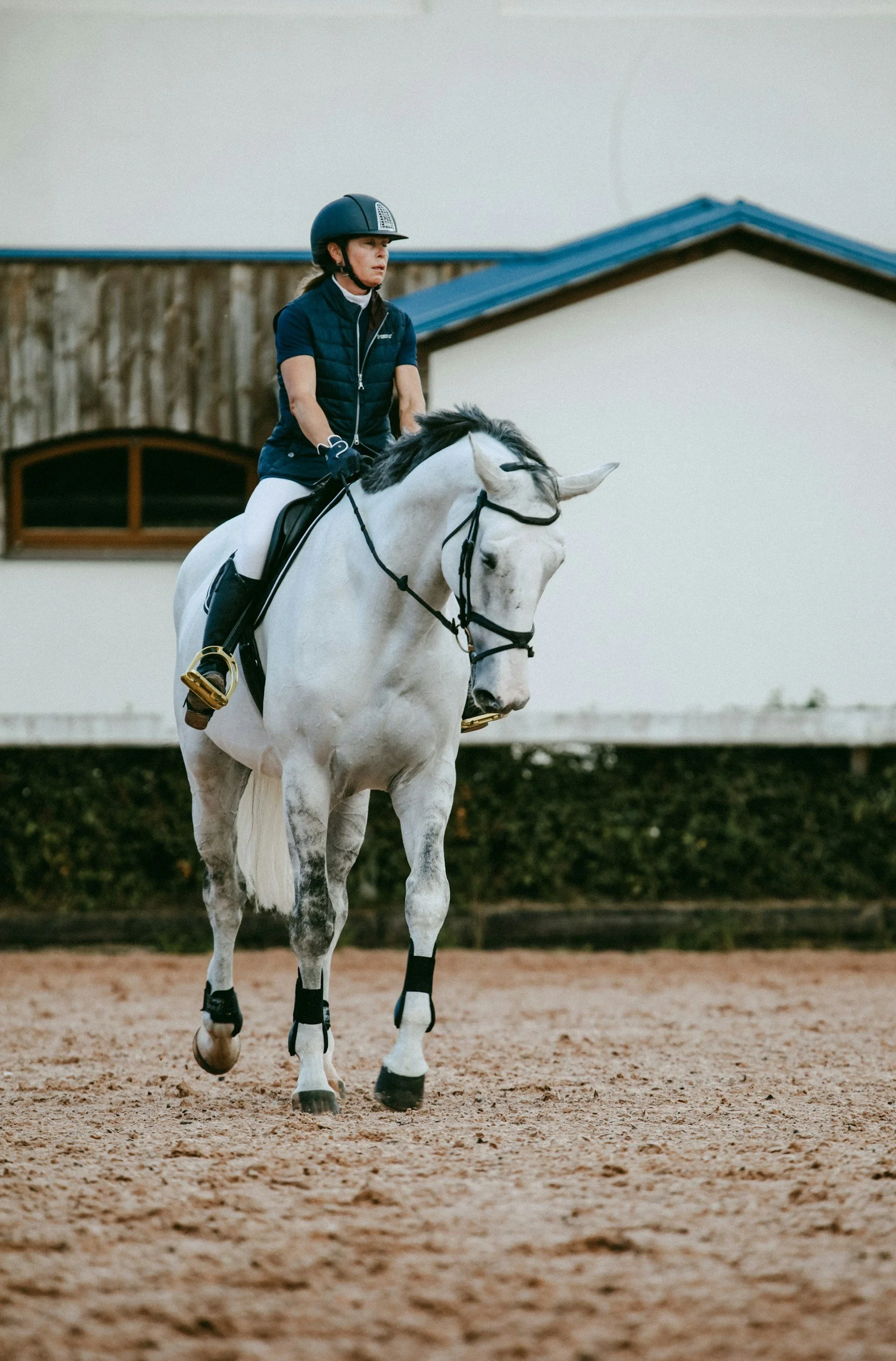 A woman riding a white horse on a sandy arena with a wooden and white building in the background.