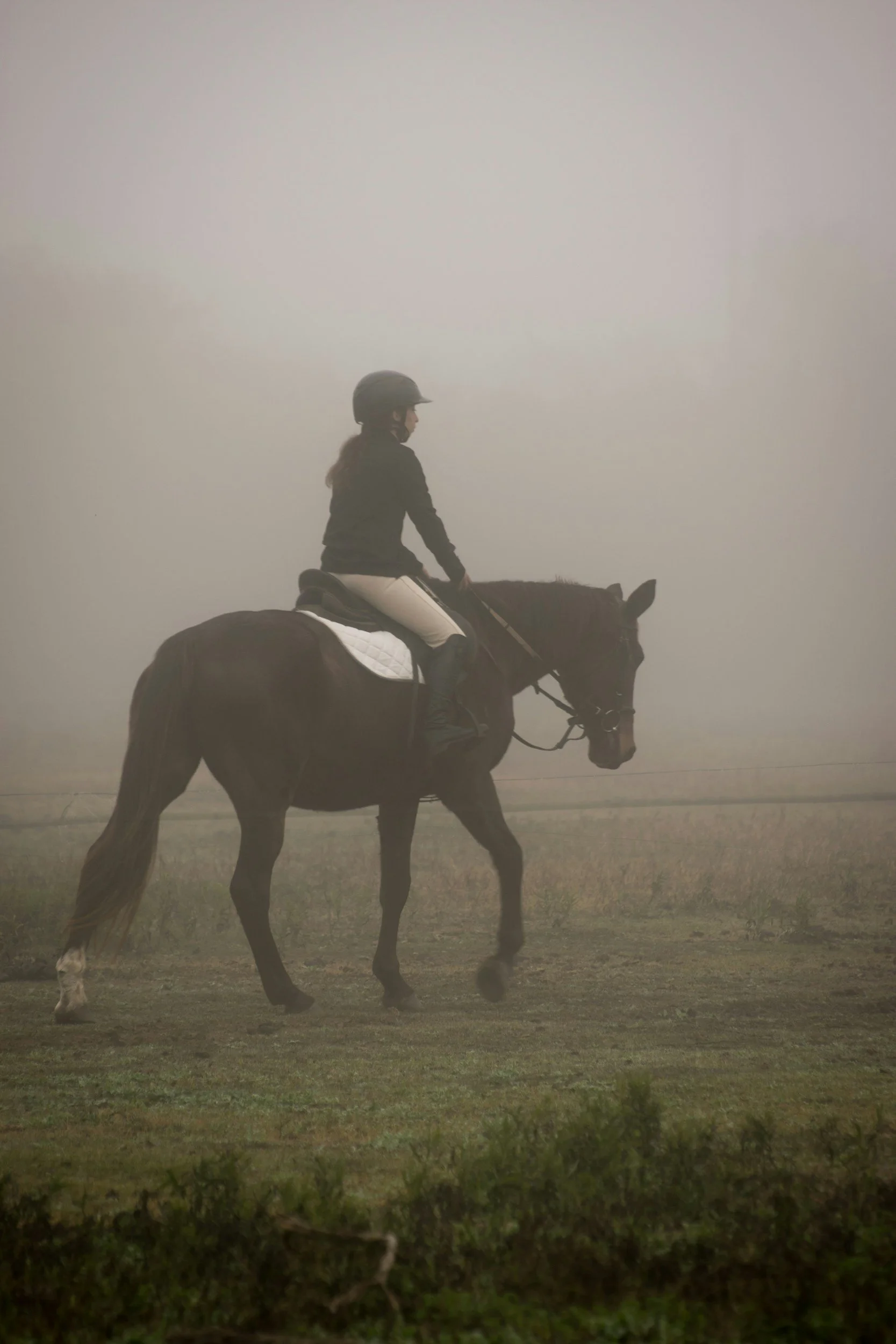 A woman wearing a helmet riding a horse in a foggy outdoor setting.