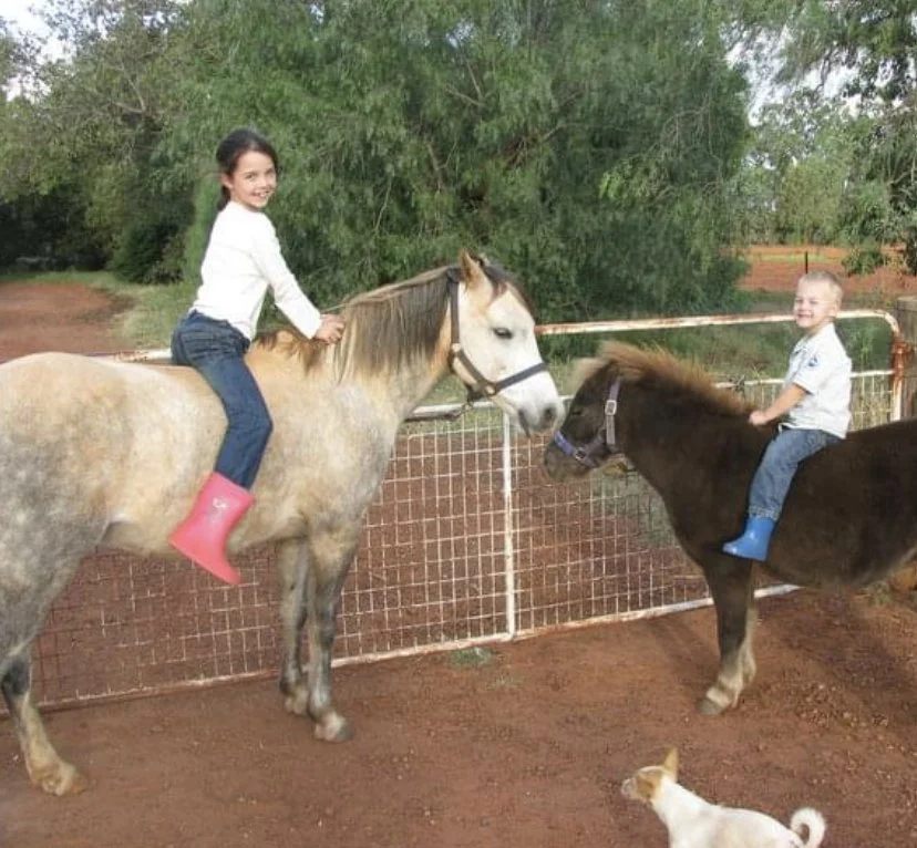 Two children riding ponies face-to-face, smiling, with a dog at their feet and greenery in the background.