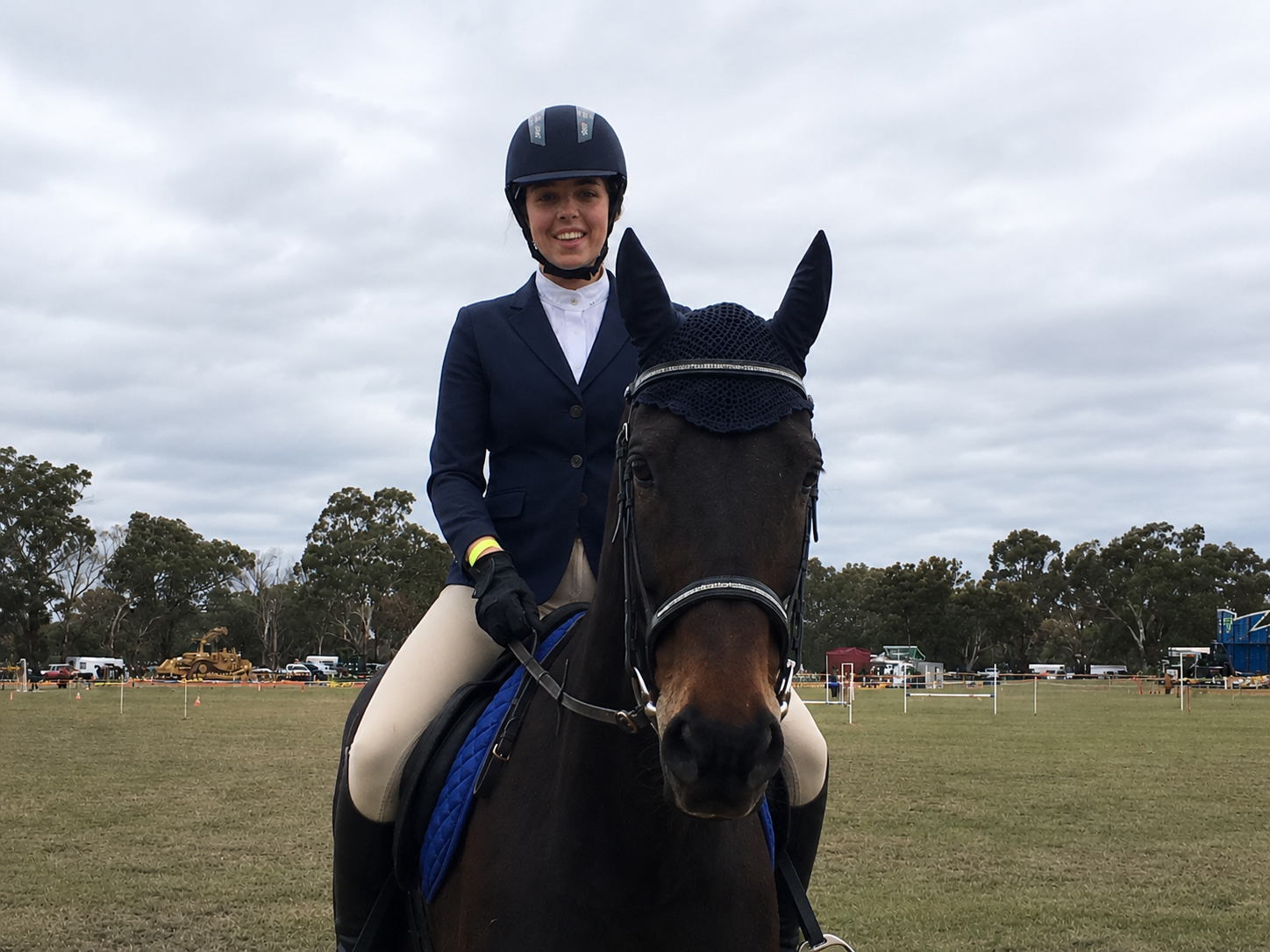 Young woman in equestrian attire riding a dark horse on a grassy field, wearing a helmet and gloves, with trees and vehicles in the background under a cloudy sky.