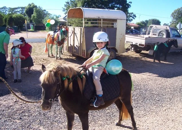 Child riding a small brown pony with a blue balloon attached to its saddle at an outdoor event.