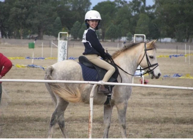 Girl riding a light gray horse in an outdoor riding arena, wearing a white helmet and navy jacket.