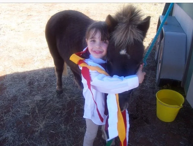 A young girl hugging a small black pony outdoors on dry grass with a yellow bucket nearby.