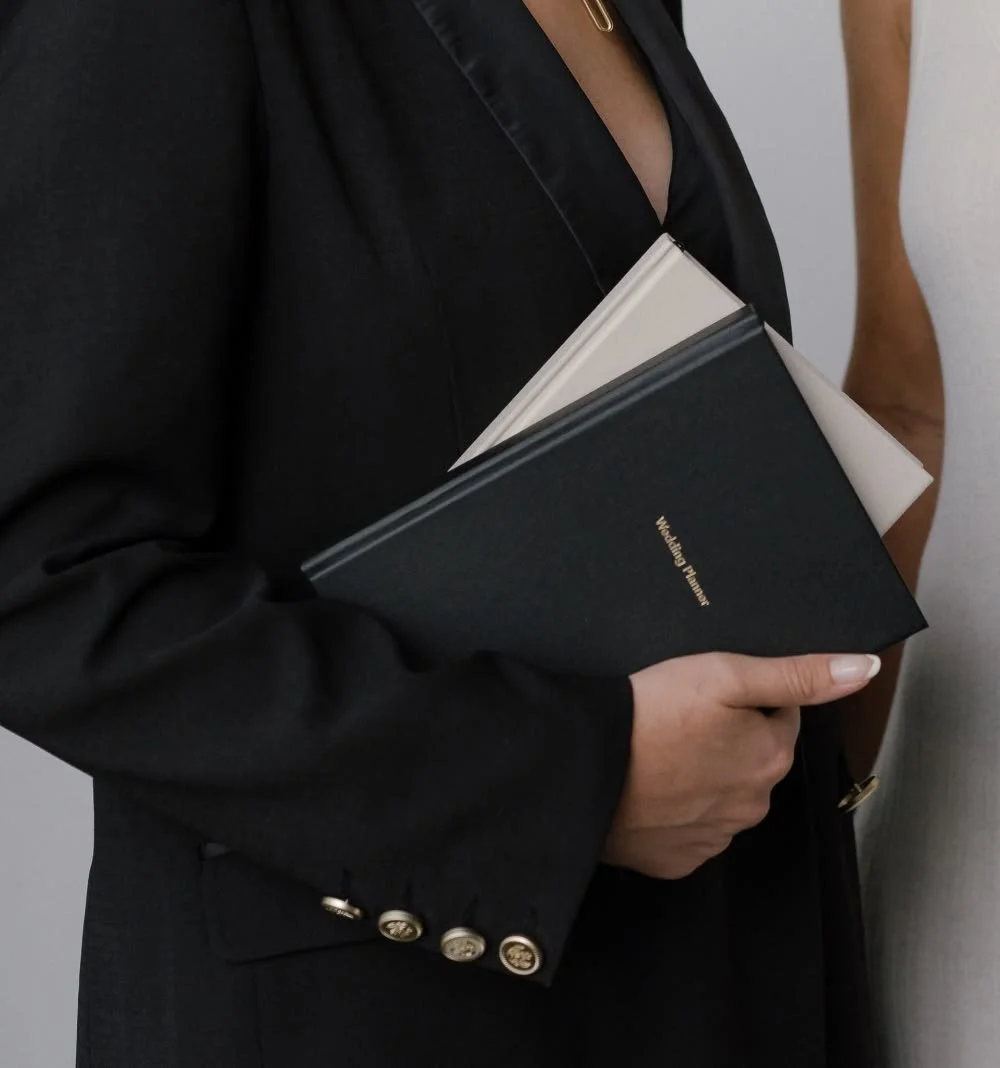 Close-up of a woman in a black blazer holding a black wedding planner and a notepad.