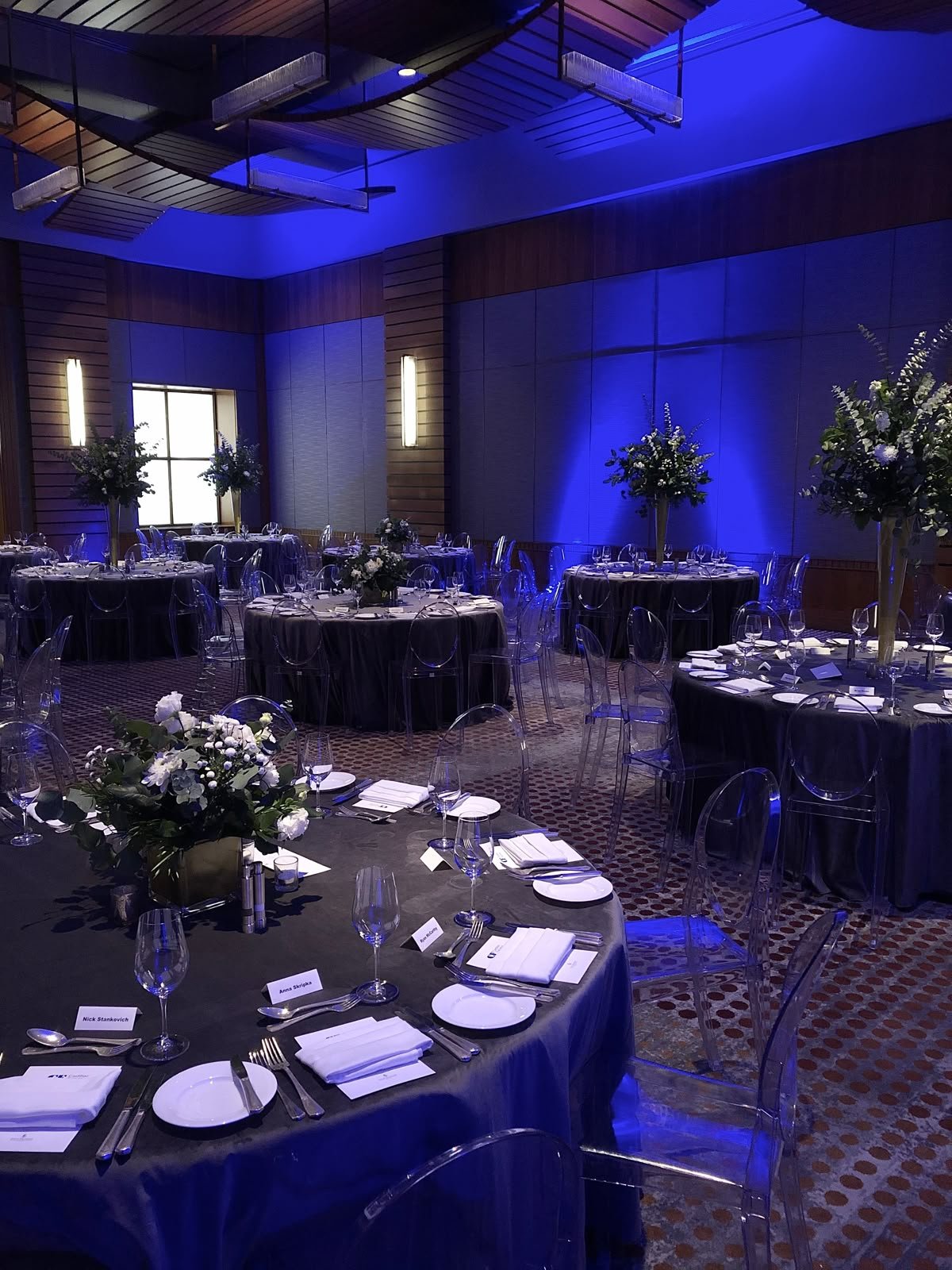 A banquet hall decorated for a formal event with round tables draped in black tablecloths, set with white plates, silverware, napkins, and glassware. Tall floral centerpieces with white flowers and greenery decorate each table. The room has wood-paneled walls, blue lighting, and modern clear chairs, creating an elegant ambiance.