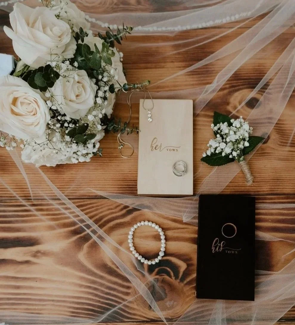 Wedding vows books for bride and groom and their rings scattered on a wooden surface with bouquet of white roses, baby's breath, greenery, and a pearl bracelet.