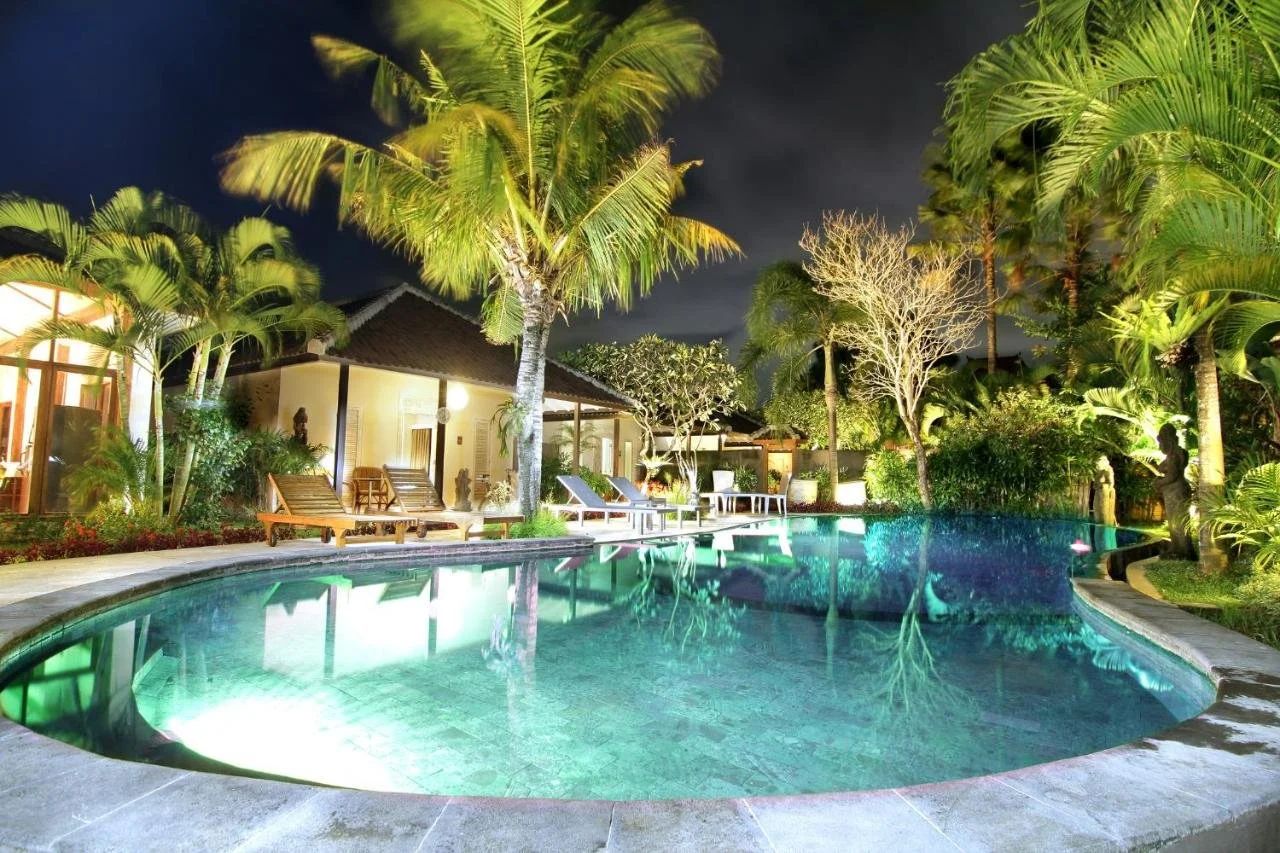 Nighttime view of a luxurious outdoor swimming pool surrounded by tropical trees and lounge chairs, with a villa in the background.