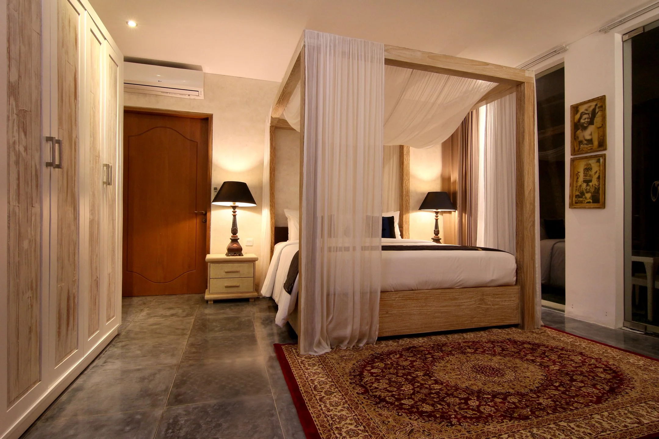 A bedroom with a four-poster bed featuring light-colored drapes, a nightstand with a black lampshade, a wooden wardrobe, framed artwork on the wall, and a patterned area rug.