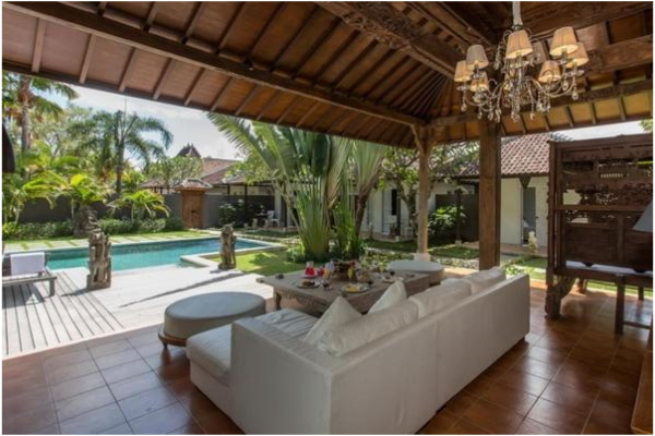 Outdoor patio with white sofa, dining table, and pool surrounded by lush tropical plants and trees, with a wooden roof and chandelier overhead.