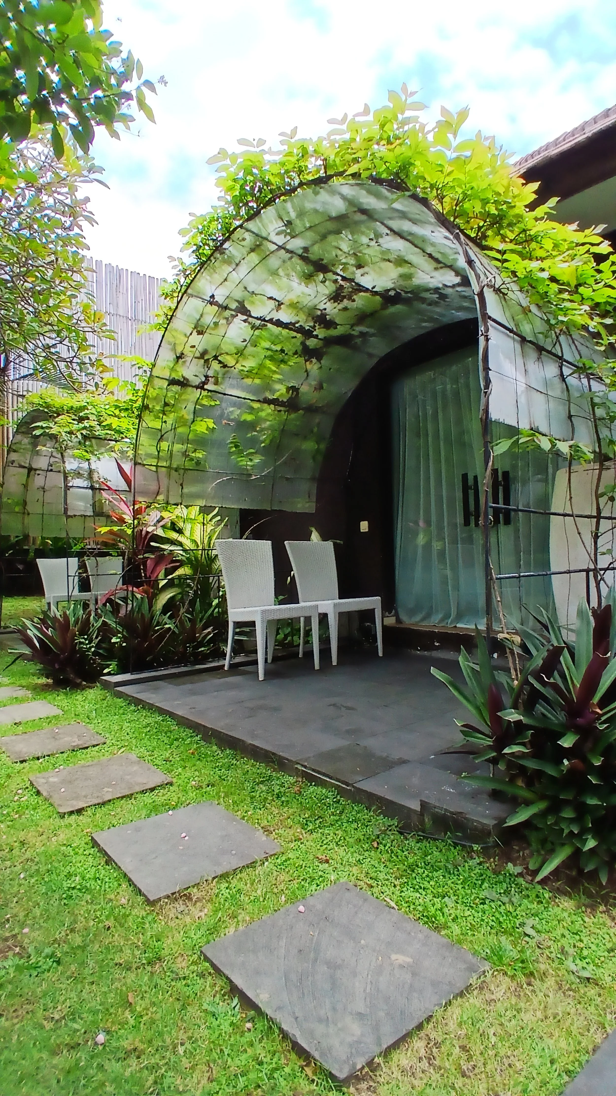 A garden with a small patio area featuring three white chairs, surrounded by lush green plants and a stone pathway leading to the patio. There is a curved, transparent greenhouse or canopy with plants growing inside or around it, and a wooden fence i