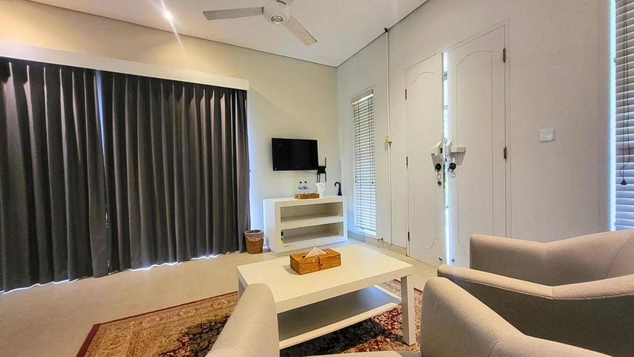 Living room with beige chairs, a white coffee table, a TV mounted on the wall, dark curtains, and a door with blinds.