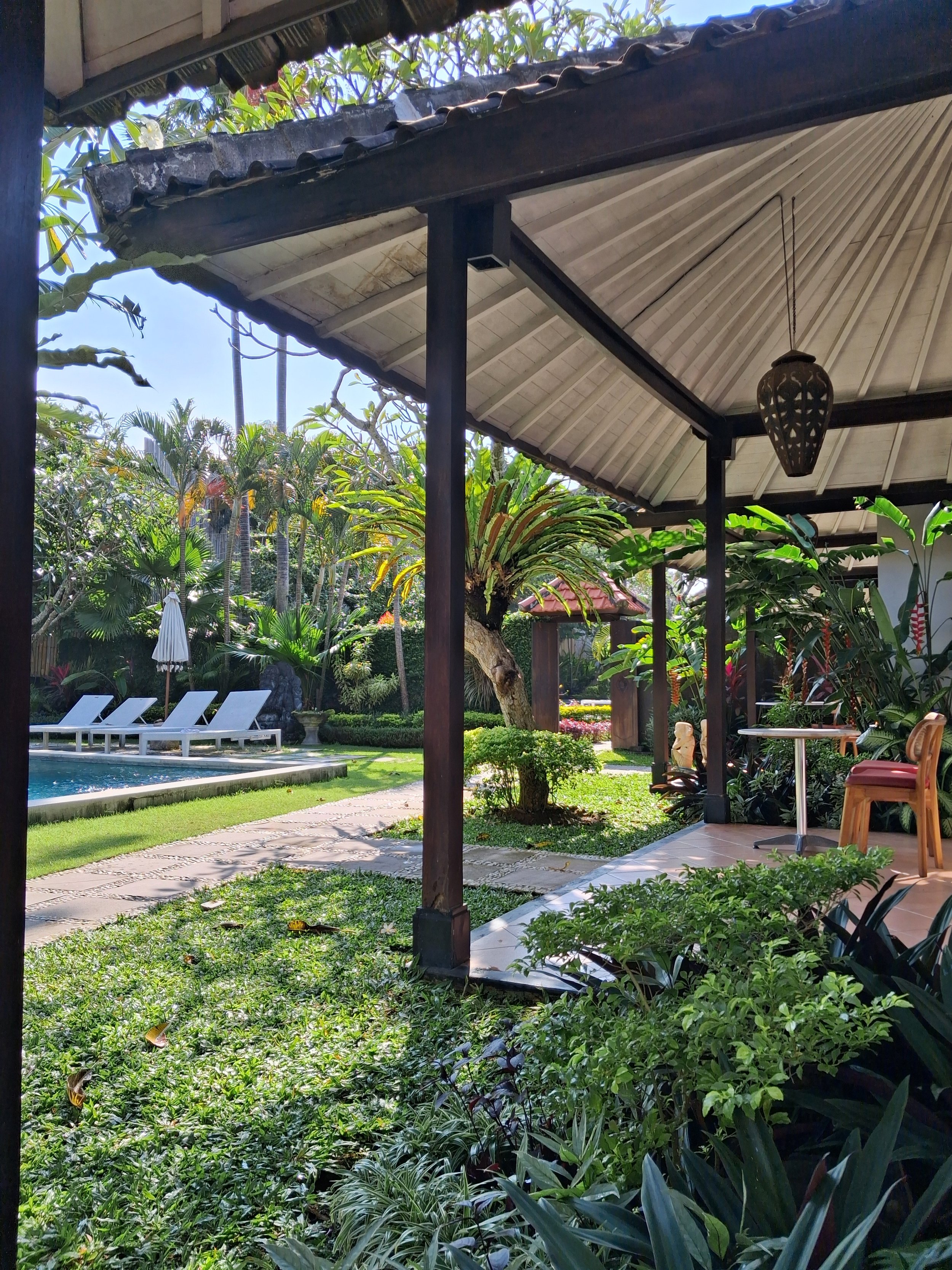 View of a lush garden with various tropical plants, a swimming pool with white lounge chairs, and a shaded seating area under a roof with a hanging lantern.