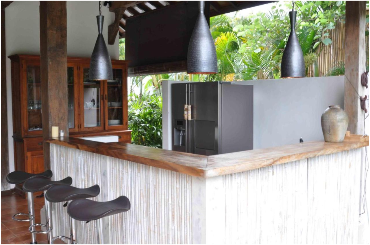 A kitchen with a wooden bar counter, black bar stools, and black hanging pendant lights. There is a gray refrigerator, wooden cabinet, a pottery vase, and lush greenery outside the windows.