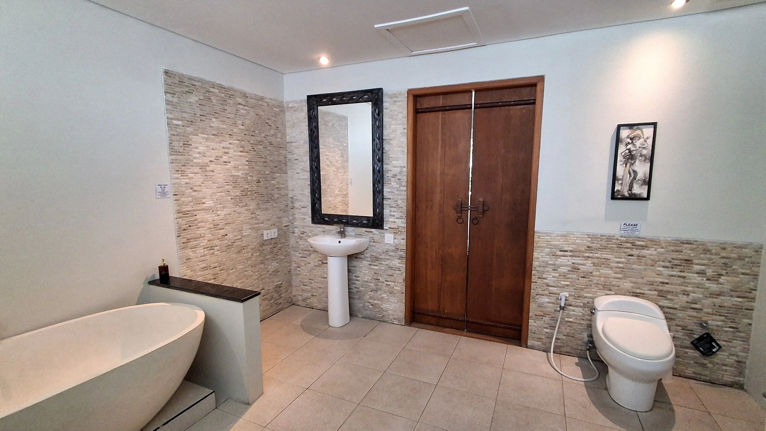 A modern bathroom with a freestanding bathtub on the left, a pedestal sink with a black mirror above it in the middle, and a toilet with bidet attachment on the right. The room features a partial tile wall with small beige stones, a wooden door with 