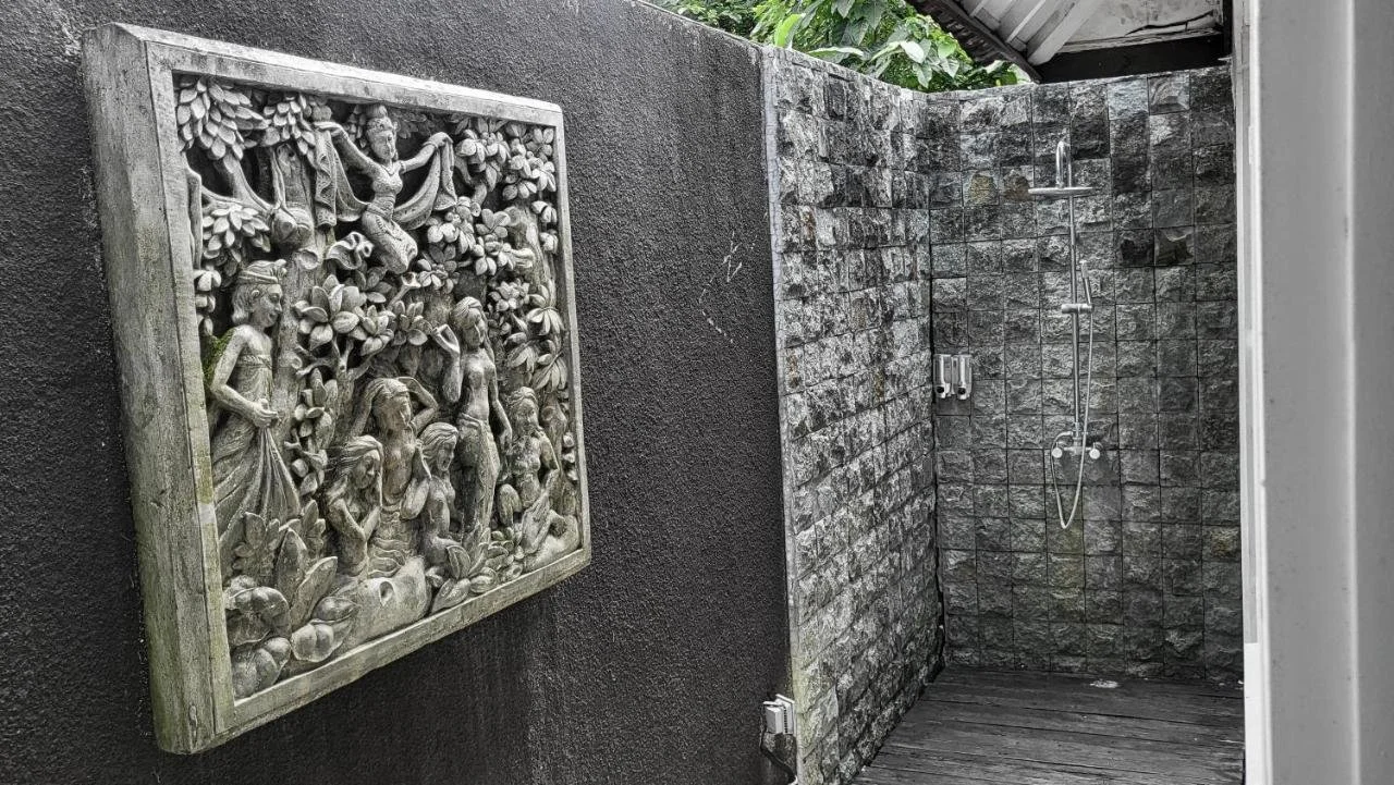 Outdoor shower with brick wall, wooden floor, and decorative stone framed carving of women in traditional dress surrounded by leaves.
