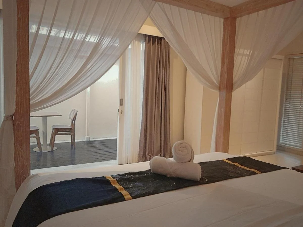 A bedroom with a four-poster bed, draped with white sheer curtains, and towels rolled on the bed. Behind the bed, there is a small balcony area with two chairs and a table, partially visible through sliding glass doors with beige curtains.