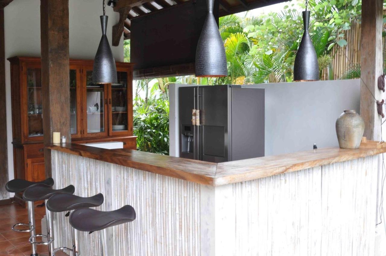A kitchen with a wooden bar counter, black bar stools, and black hanging pendant lights. There is a gray refrigerator, wooden cabinet, a pottery vase, and lush greenery outside the windows.