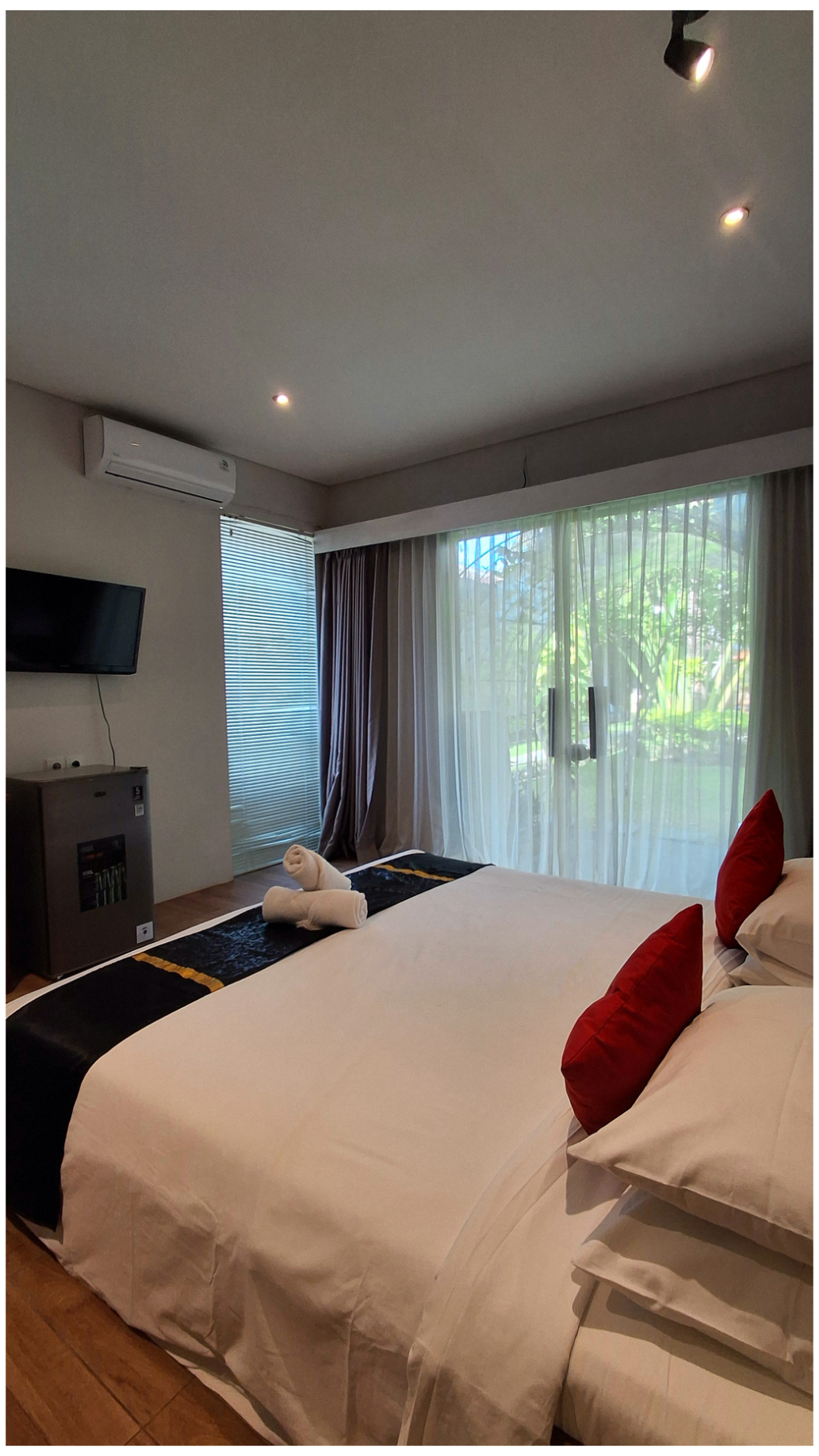 A room with a bed, red pillows, white sheets, rolled towels, a flat-screen TV on the wall, window with blinds and sheer curtains, and a view of greenery outside.