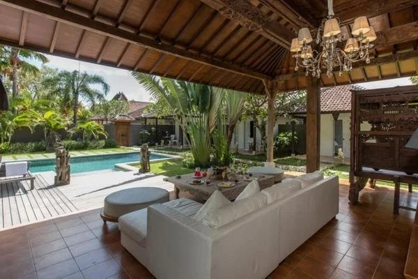 Outdoor patio with white sofa, dining table, and pool surrounded by lush tropical plants and trees, with a wooden roof and chandelier overhead.