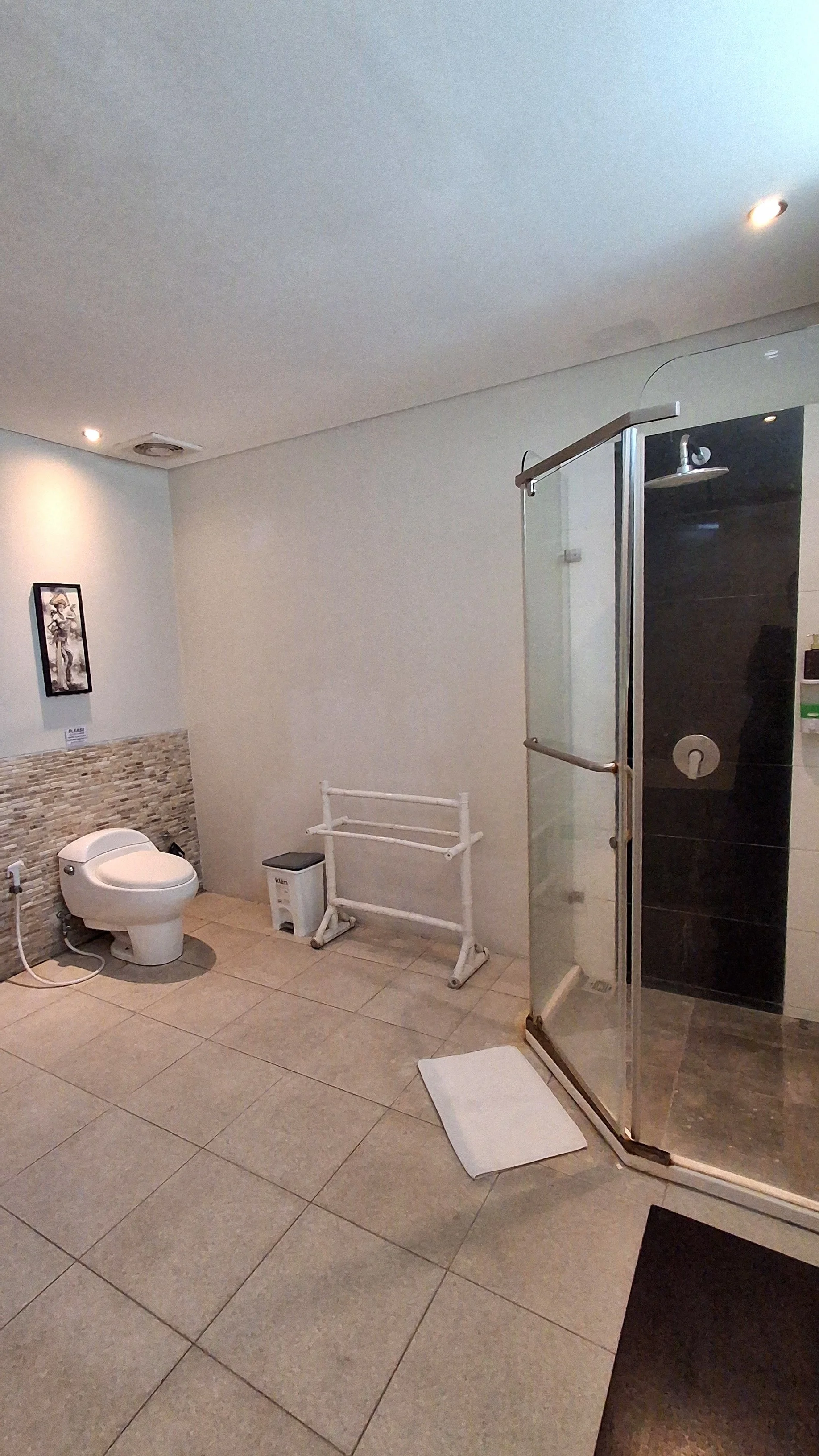 A bathroom with a toilet, a small trash bin, a white towel rack, a shower with clear glass door, and a framed picture on the wall.