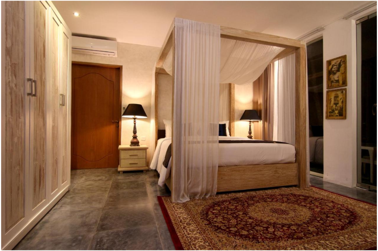 A bedroom with a four-poster bed featuring light-colored drapes, a nightstand with a black lampshade, a wooden wardrobe, framed artwork on the wall, and a patterned area rug.