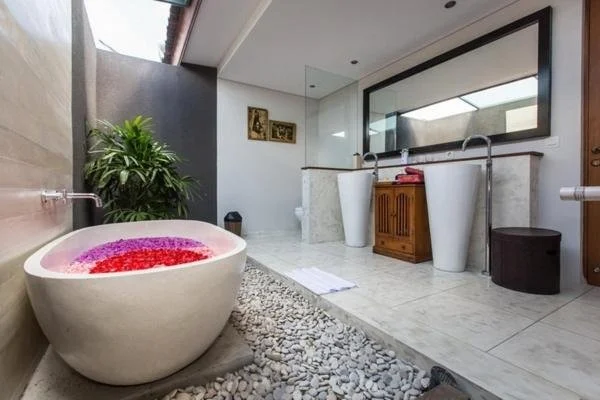 Bathroom with stone tub filled with pink and red flower petals, pebbled walkway, two white pedestals with faucets, wooden storage cabinet, large mirror, and potted plant.