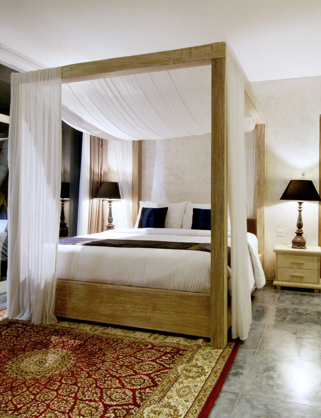 Decorative bedroom with four-poster bed with white curtains, white bedding, dark pillows, two bedside tables with black lamps, and a red and beige patterned rug on gray tiled floor.