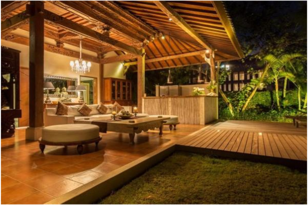 Exterior view of a covered outdoor patio at night with wooden flooring, a chandelier, and a seating area with cushioned furniture, overlooking a lush garden.