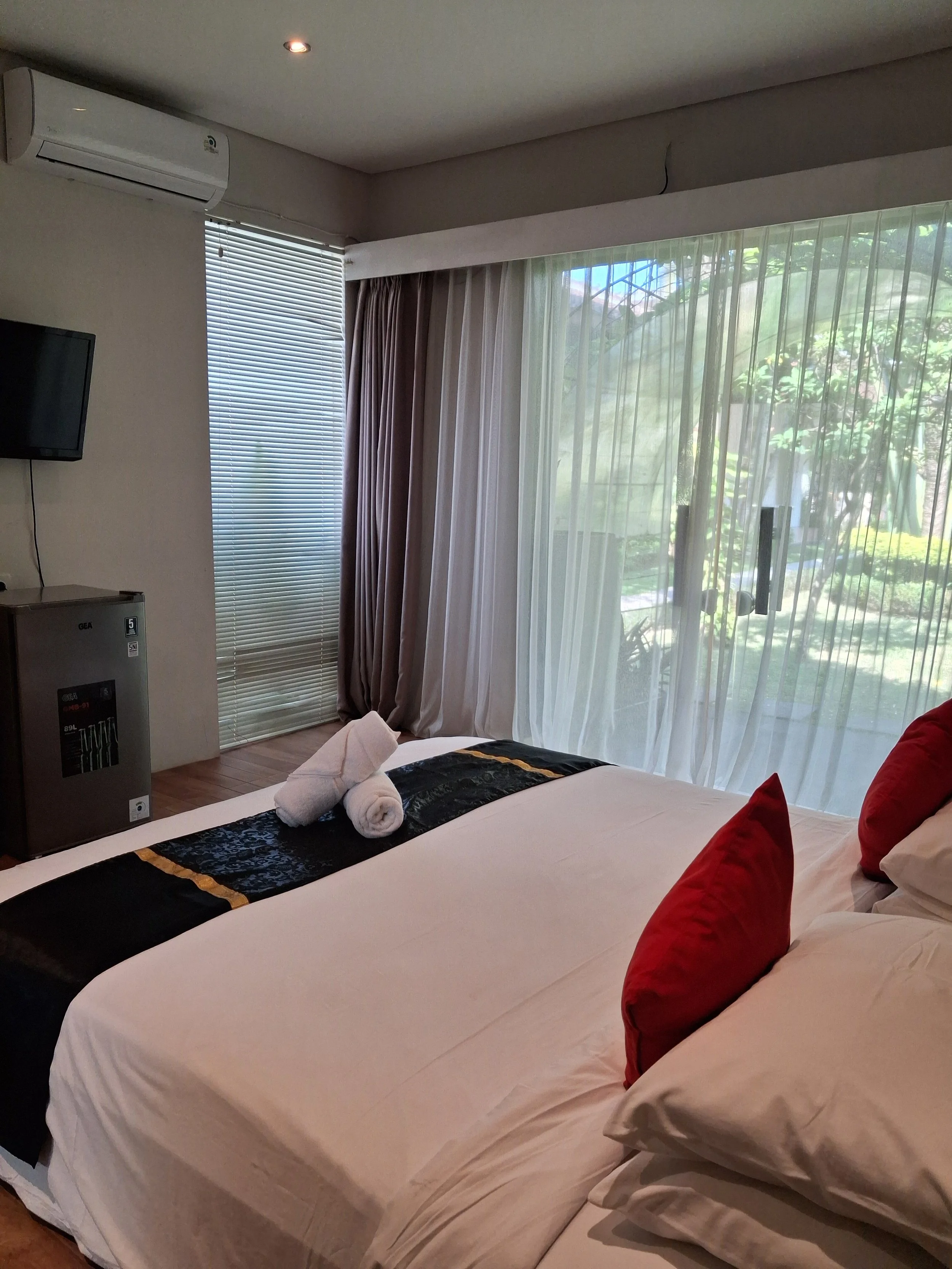 A hotel room with a bed, red pillows, white sheets, rolled towels, a flat-screen TV on the wall, window with blinds and sheer curtains, and a view of greenery outside.