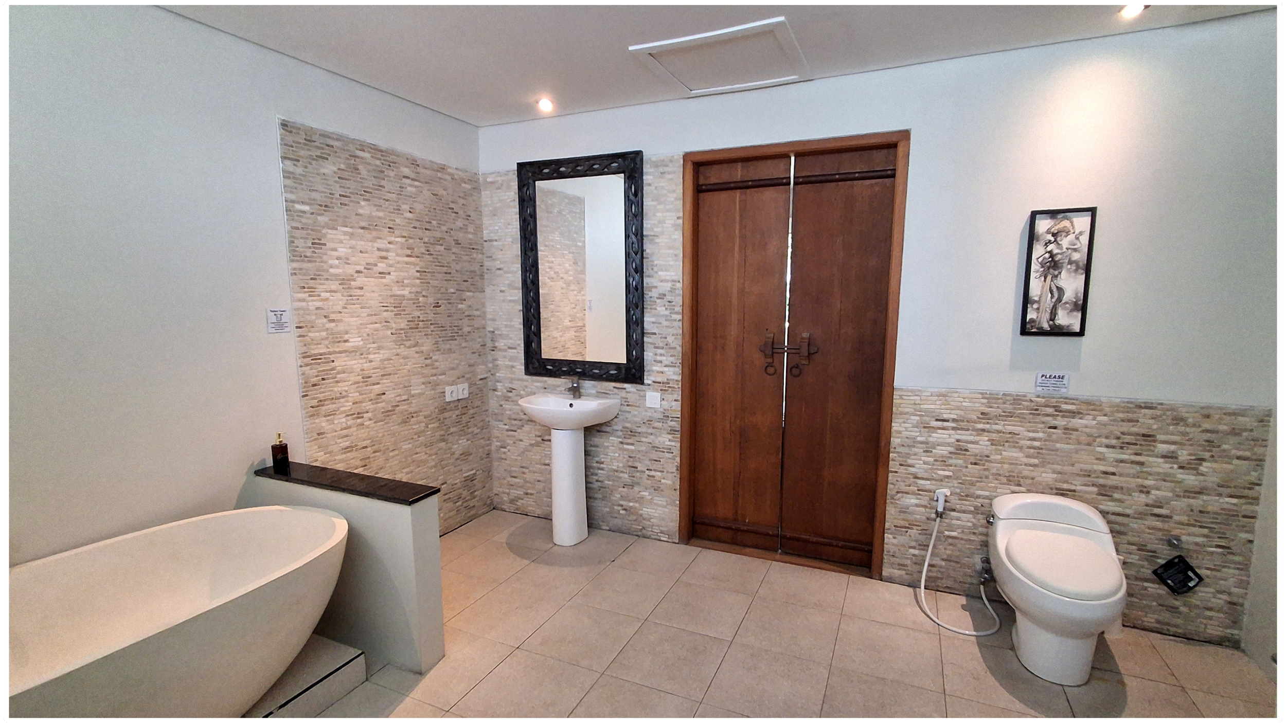 A modern bathroom with a freestanding bathtub on the left, a pedestal sink with a black mirror above it in the middle, and a toilet with bidet attachment on the right. The room features a partial tile wall with small beige stones, a wooden door with 