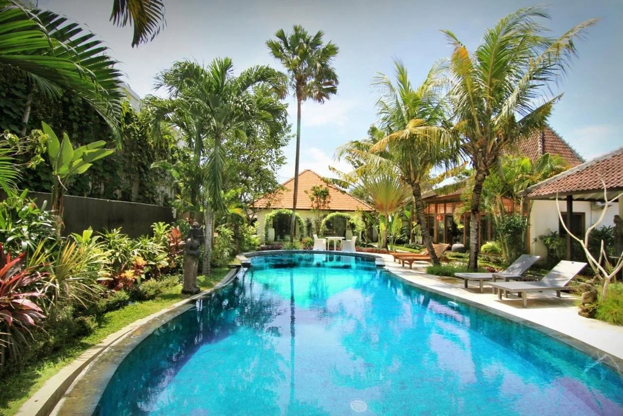 Tropical poolside scene with clear blue swimming pool, lush green palm trees, lounge chairs, and bungalows with tiled roofs in the background.