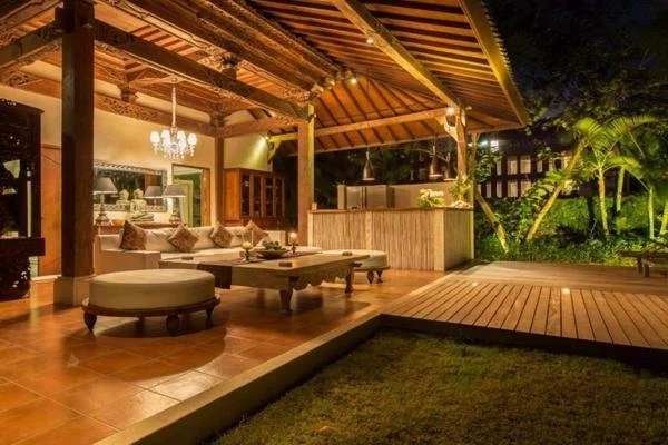 Exterior view of a covered outdoor patio at night with wooden flooring, a chandelier, and a seating area with cushioned furniture, overlooking a lush garden.