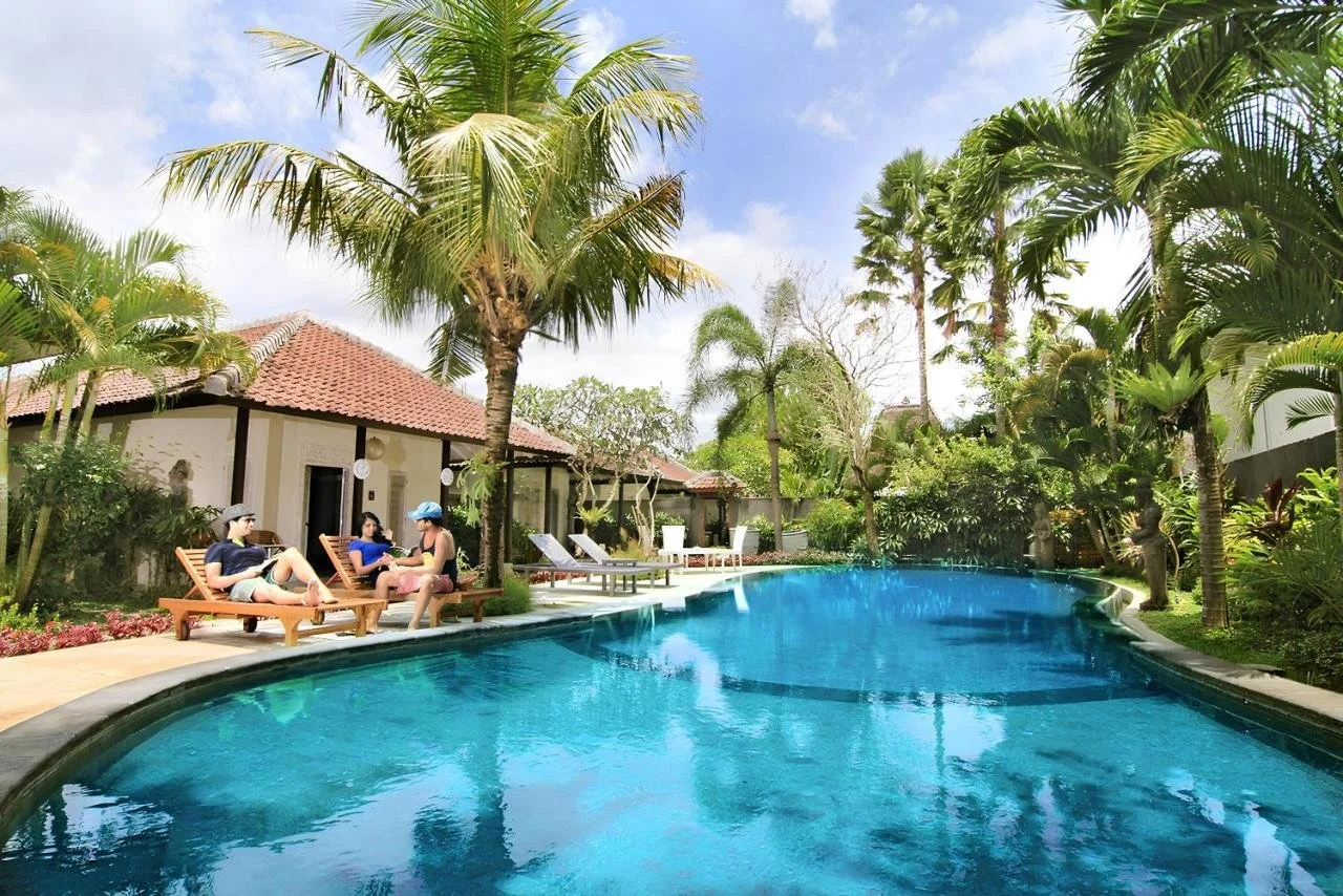 A tropical pool area with lounge chairs, palm trees, and a clear blue swimming pool surrounded by lush greenery and a small building.