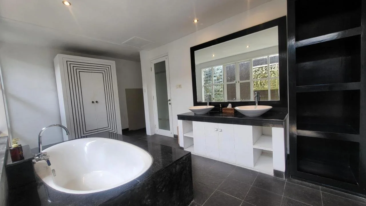 Modern bathroom with black and white color scheme, double vanity with vessel sinks, large mirror, black shelving unit, bathtub, and window overlooking greenery.