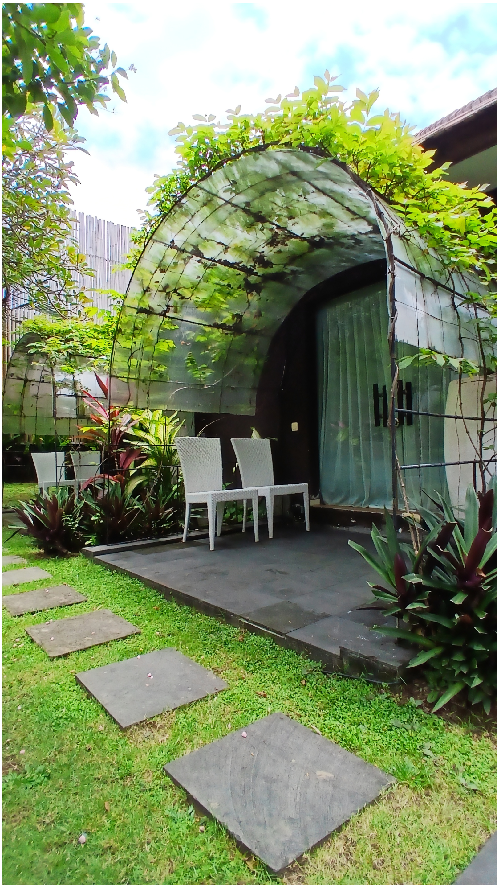 A garden with a small patio area featuring three white chairs, surrounded by lush green plants and a stone pathway leading to the patio. There is a curved, transparent greenhouse or canopy with plants growing inside or around it, and a wooden fence i