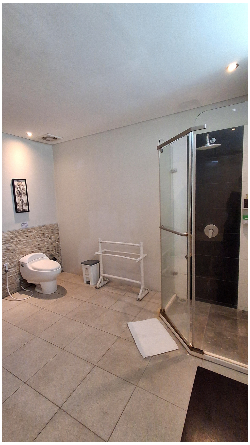 A bathroom with a toilet, a small trash bin, a white towel rack, a shower with clear glass door, and a framed picture on the wall.
