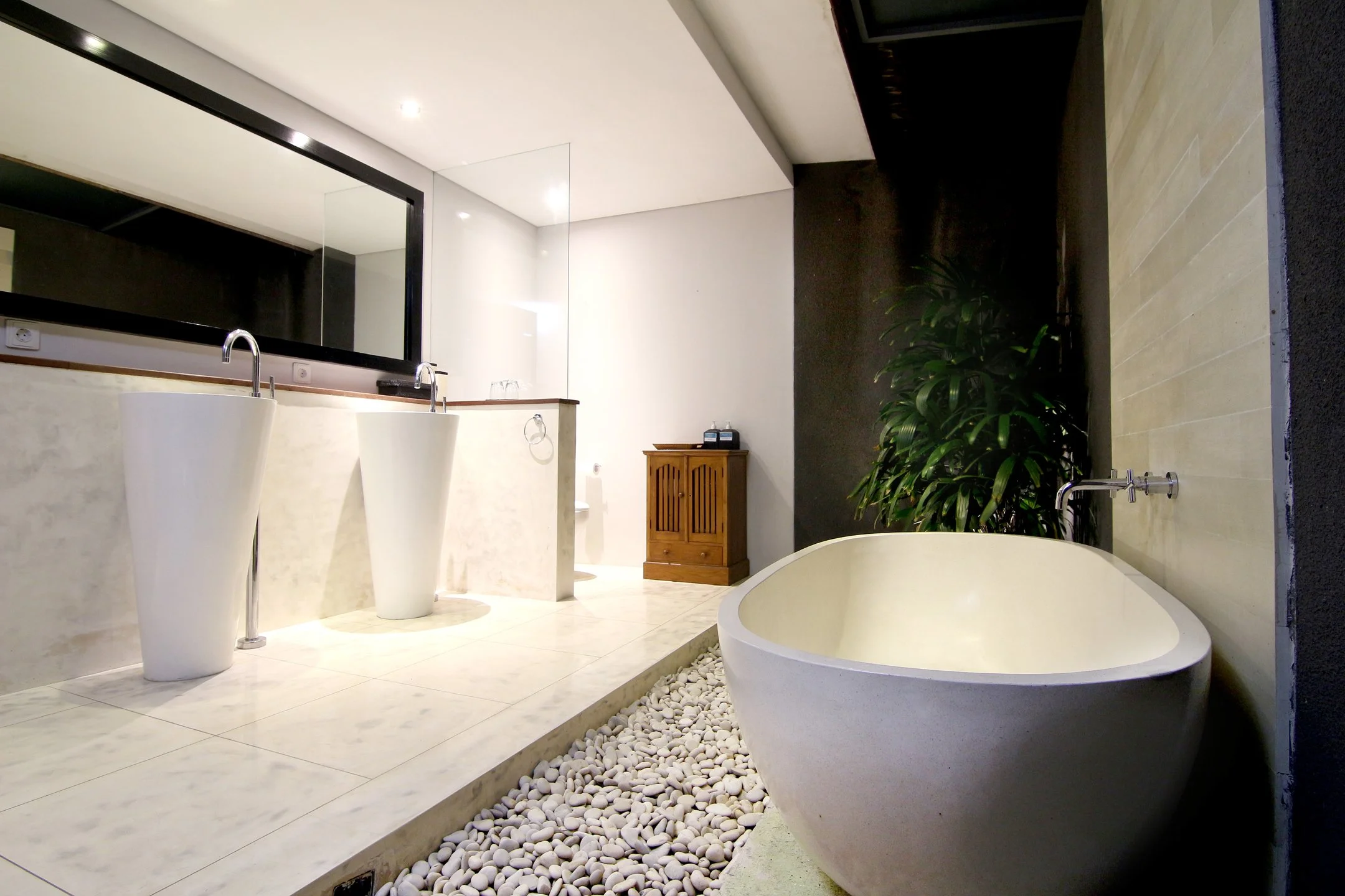 Modern bathroom with a freestanding bathtub, two pedestal sinks with faucets, a large mirror, white pebbles along the edge of the tub, a small wooden cabinet, and lush green plants, with beige tiled flooring and a black wall.