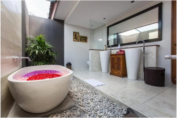 Bathroom with stone tub filled with pink and red flower petals, pebbled walkway, two white pedestals with faucets, wooden storage cabinet, large mirror, and potted plant.