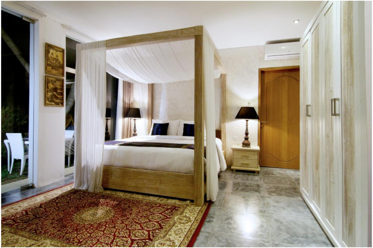 Decorative bedroom with four-poster bed with white curtains, white bedding, dark pillows, two bedside tables with black lamps, and a red and beige patterned rug on gray tiled floor.