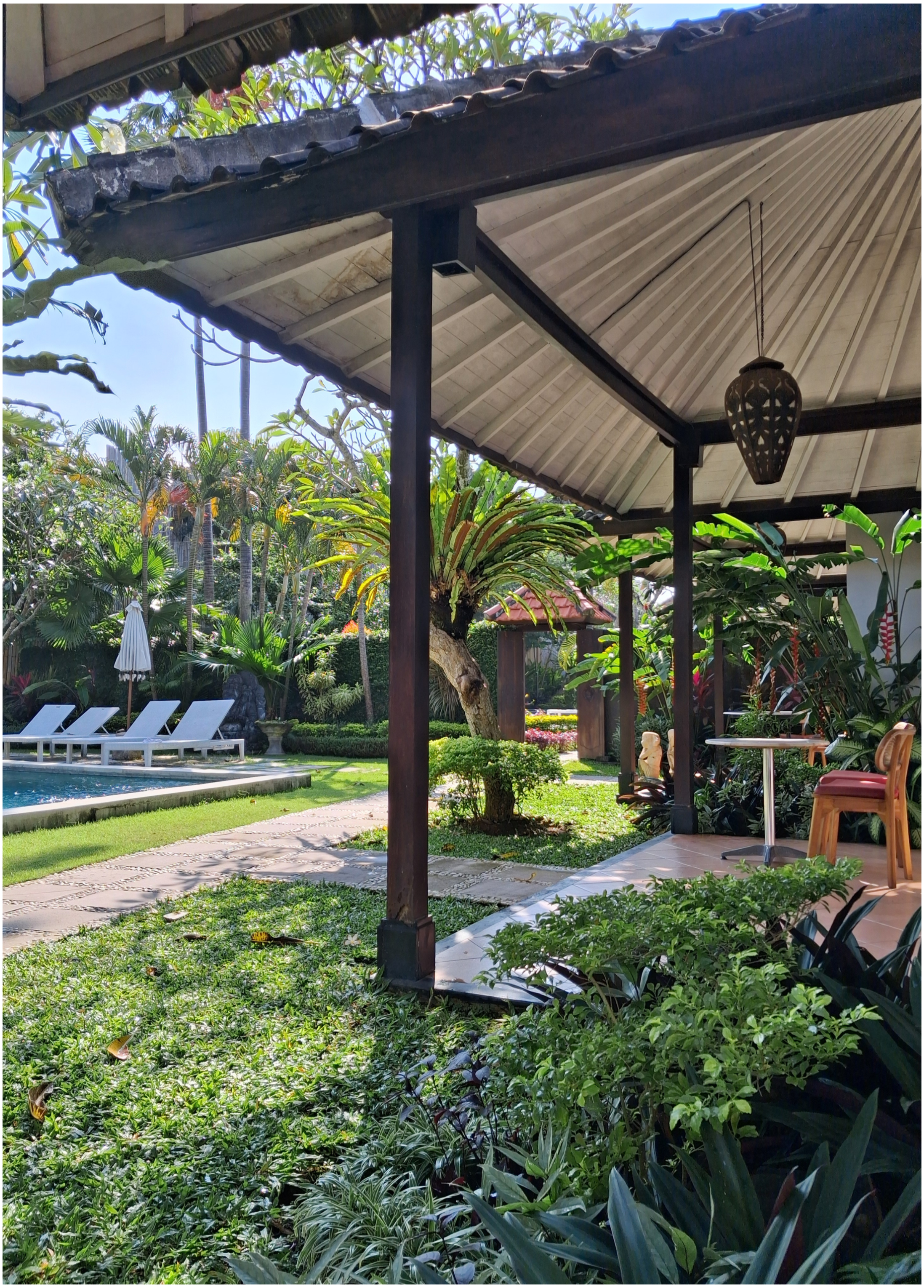 View of a lush garden with various tropical plants, a swimming pool with white lounge chairs, and a shaded seating area under a roof with a hanging lantern.